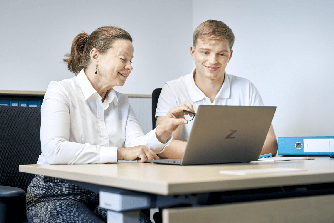 Ein junger Mann und eine Frau sitzen an einem Schreibtisch und schauen gemeinsam auf einen Laptop.