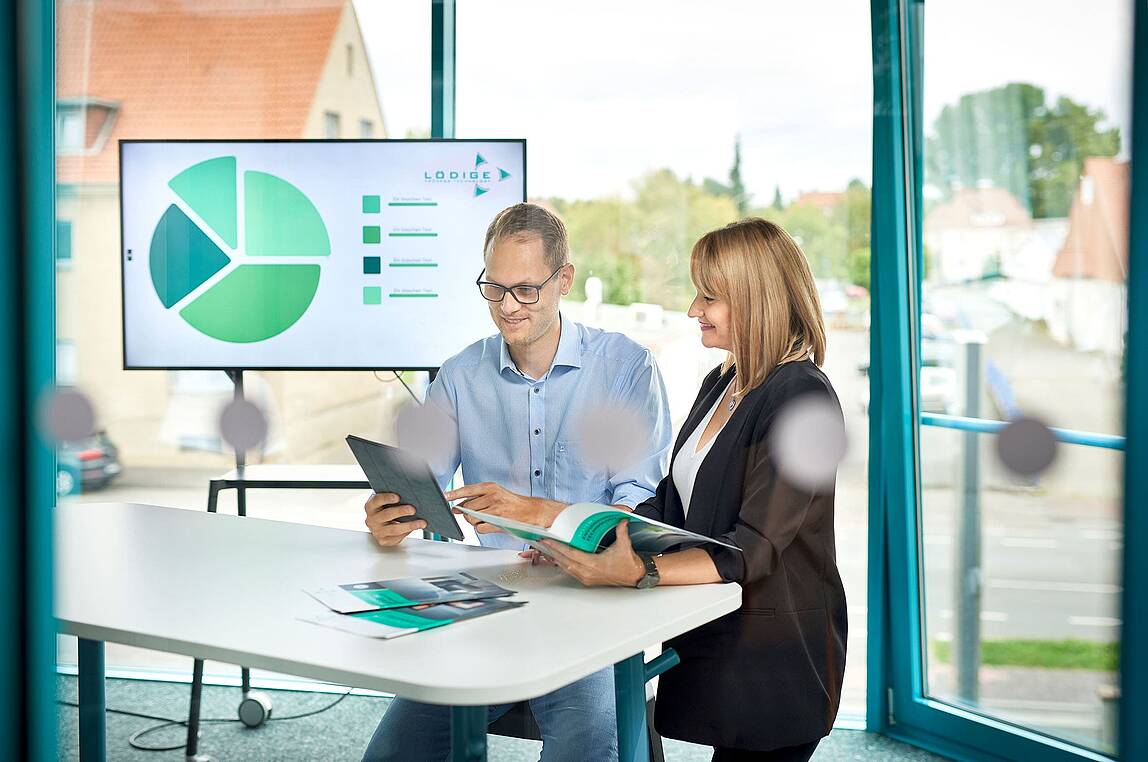 Zwei Lödige-Mitarbeiter*innen stehen in einem modernen Besprechungsraum und schauen gemeinsam auf ein Tablet.