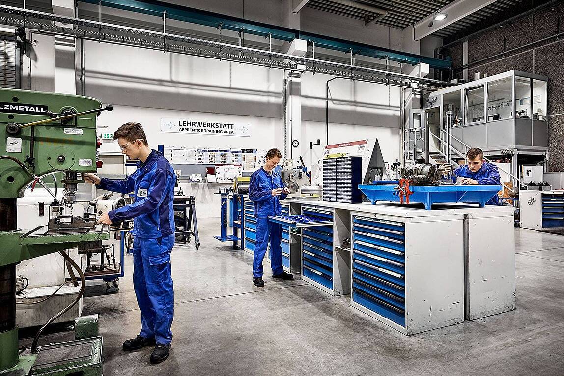 Drei Auszubildende in blauen Overalls arbeiten an verschiedenen Maschinen und Werkbänken in einer Werkstatt.