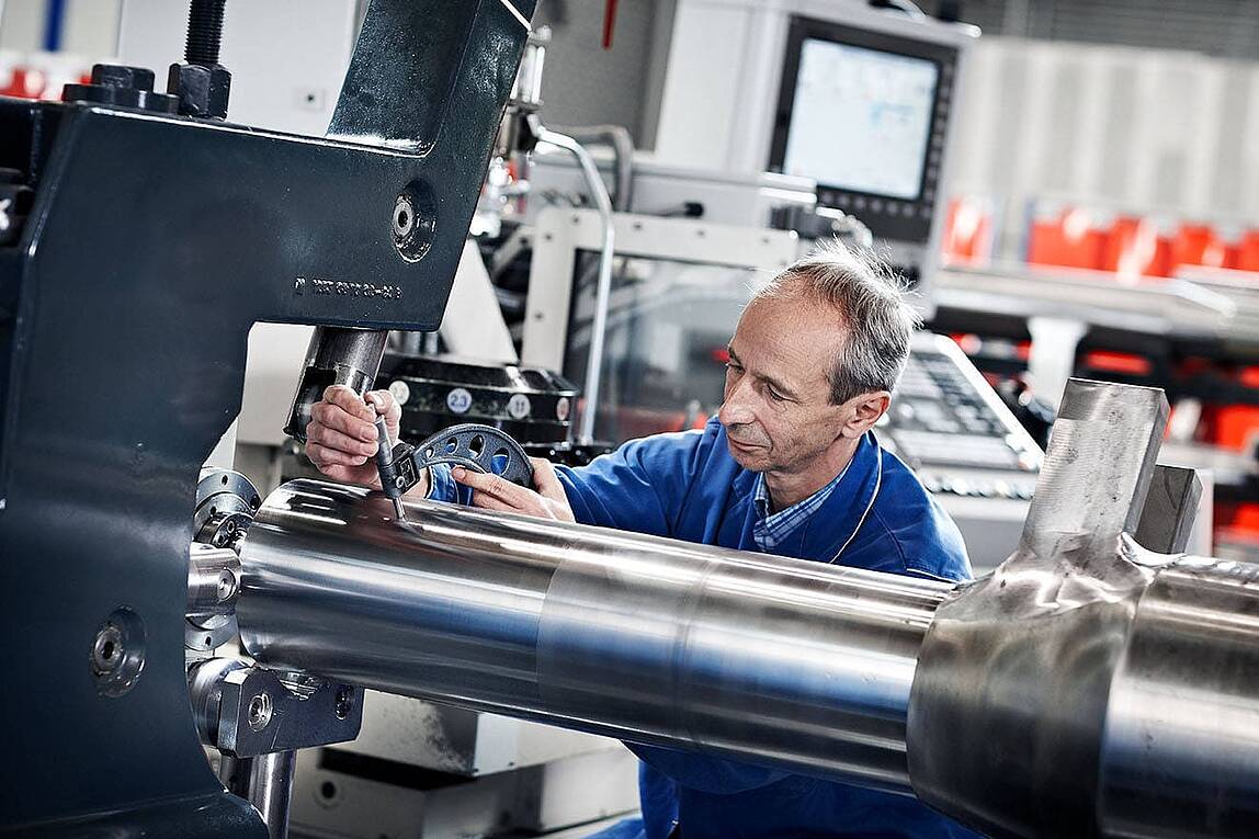 Facharbeiter im blauen Overall misst präzise eine Metallwelle an einer Industriemaschine in einer modernen Fertigungshalle.
