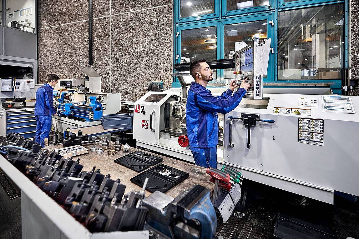 Zwei junge Zerspanungsmechaniker in blauen Overalls arbeiten an CNC-Drehmaschinen in einer großen Industriefertigungshalle.