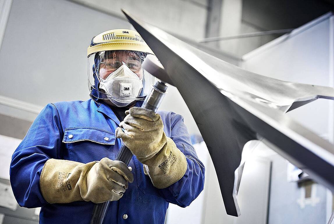 Mitarbeiter mit Atemschutzmaske, Gesichtsschutz und Schutzhandschuhen bearbeitet eine große Metallkomponente mit scharfen Kanten in einer Industriehalle.