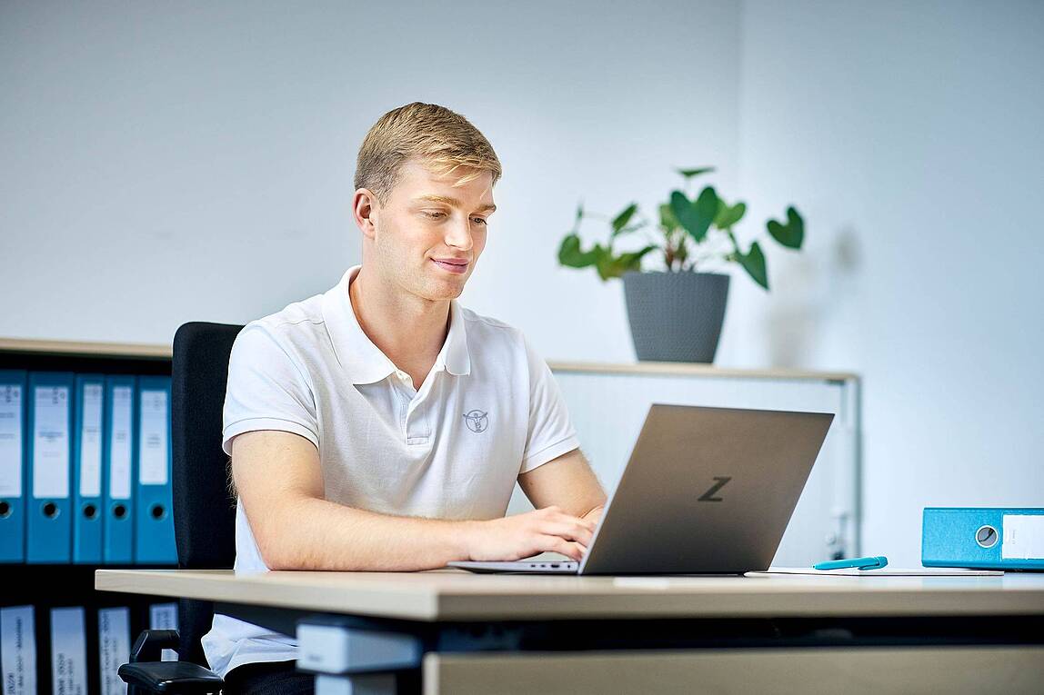 Ein Auszubildender sitzt in einem Büro an einem Schreibtisch und arbeitet an einem Laptop.