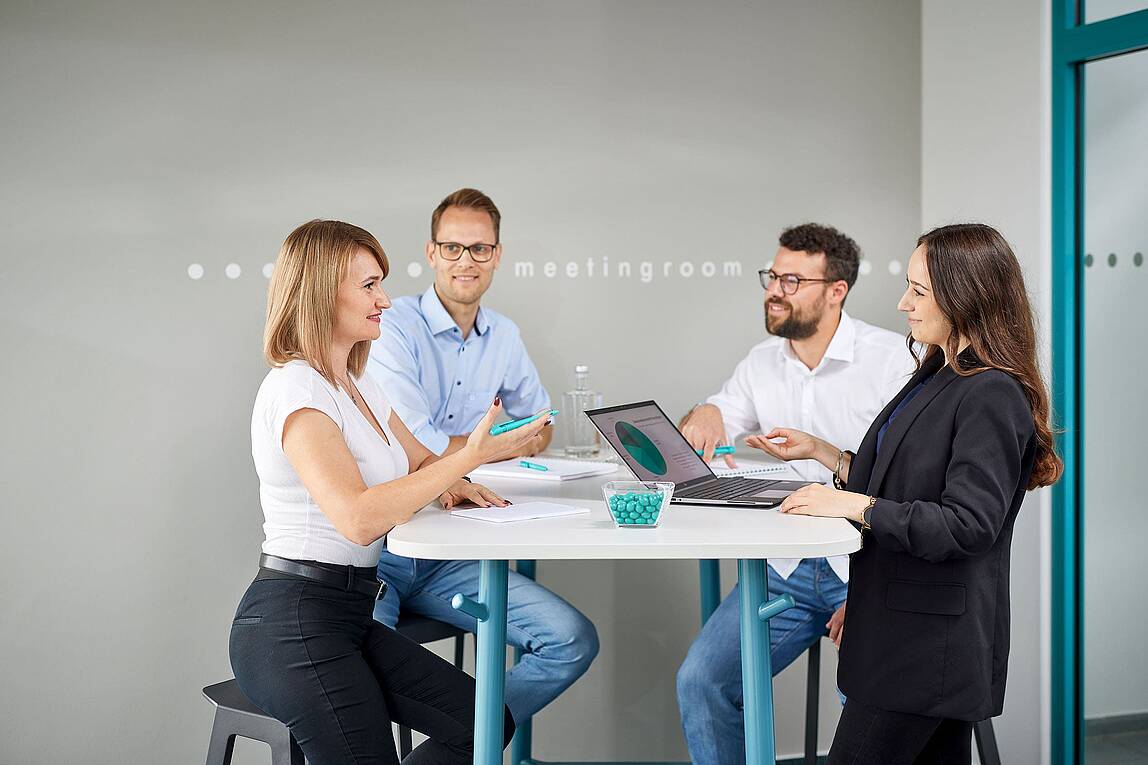 Vier Lödige-Mitarbeiter*innen stehen in einem Besprechungsraum um einen Tisch, auf dem ein Laptop steht und diskutieren.