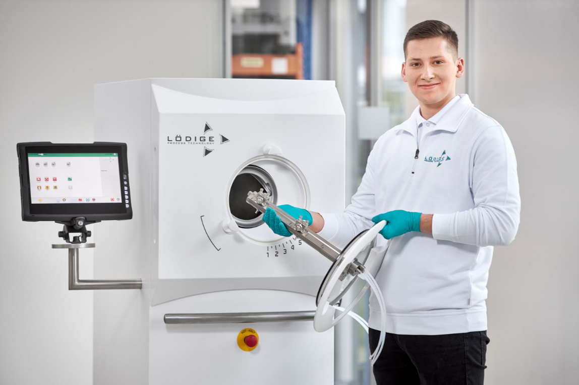 Ein junger Lödige-Mitarbeiter mit türkisen Schutzhandschuhen bedient eine weiße Prozessmaschine in einem hellen Labor.