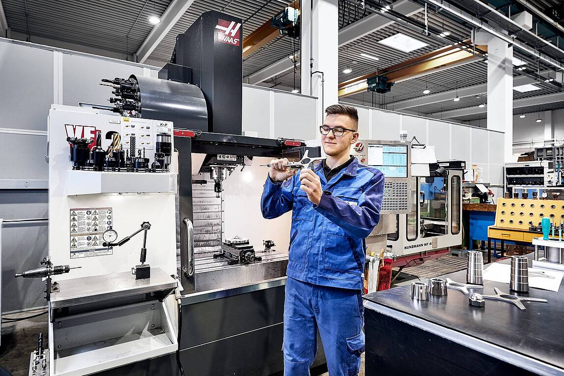 Ein junger Industriemechaniker in einem blauen Overall arbeitet an einer Maschine in einer Industriehalle.