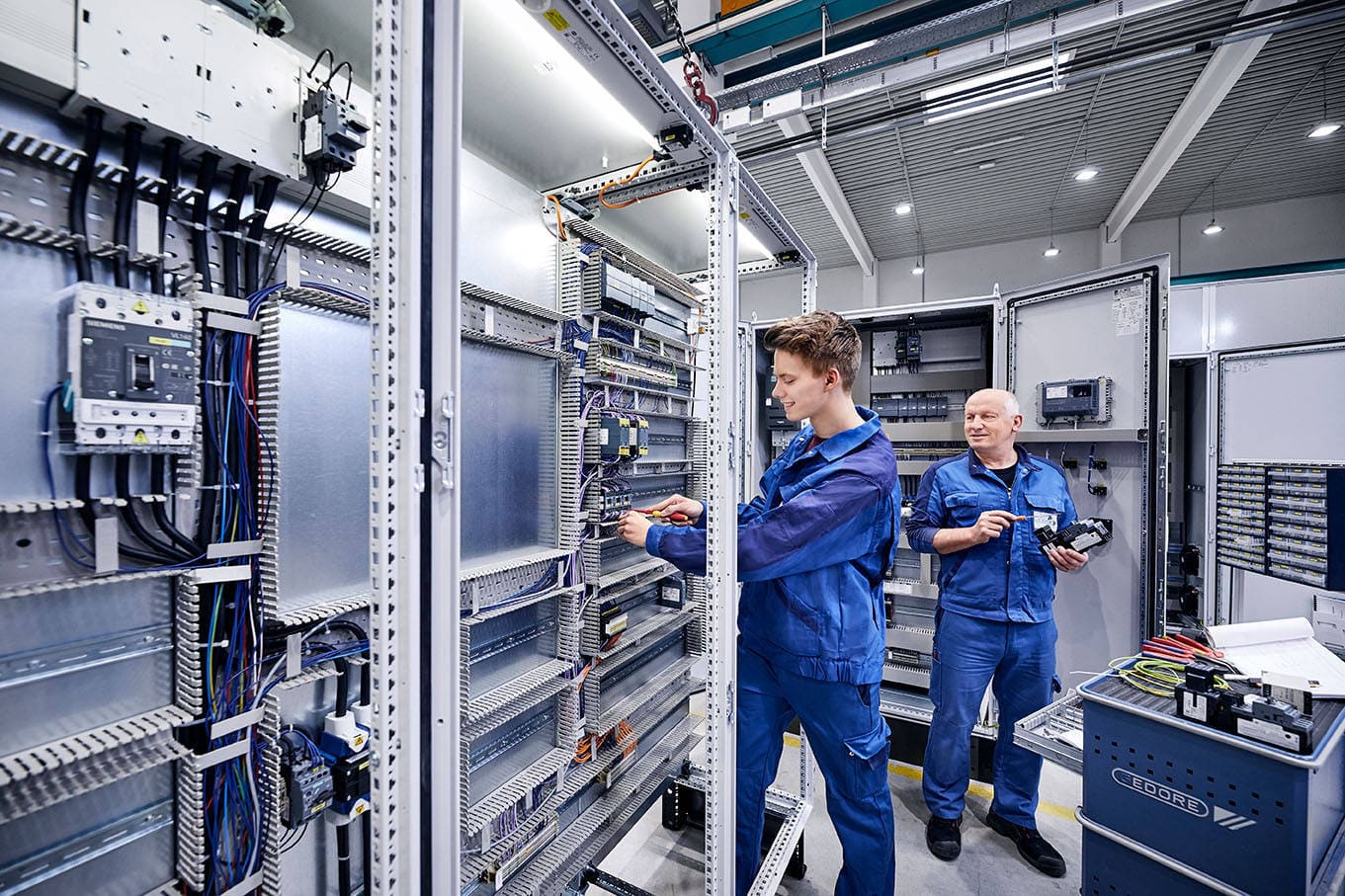 Zwei Elektriker in blauen Overalls arbeiten an einem großen Schaltschrank in einem modernen Elektrotechnik-Labor.