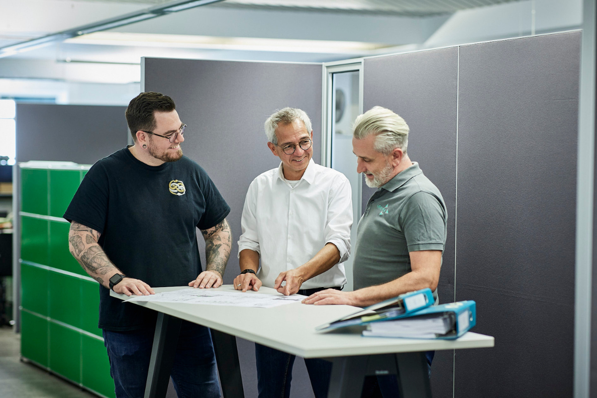 Drei Männer stehen um einen Tisch, auf dem ein Plan liegt und unterhalten sich darüber.